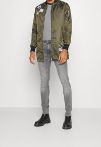 Hombre vistiendo una chaqueta bomber larga de color verde oliva con parches, camiseta a rayas en blanco y negro, jeans skinny grises y botas de cuero negras.