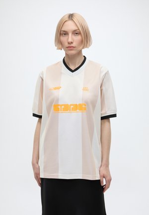 Jeune personne aux cheveux blonds courts portant un maillot de sport beige et blanc rayé avec un texte orange, debout devant un fond clair uni.