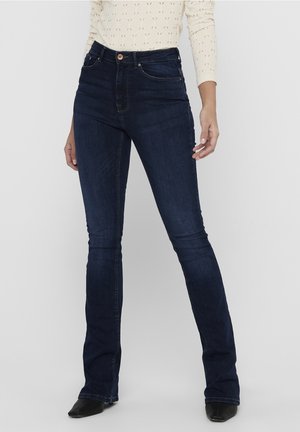 Kobieta w ciemnoniebieskich dżinsach bootcut z wysokim stanem i jasnej bluzce z długim rękawem, stojąca na jednolitym tle.