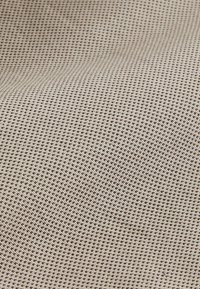 Tekstil i lys beige med et lille mønster i mørkebrun, der fremviser en glat overflade med subtil dimensionalitet.