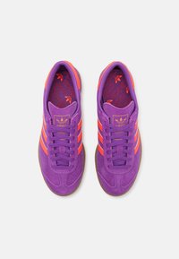 Para fioletowych zamszowych butów sportowych Adidas z pomarańczowymi paskami i logotypami, brązowymi podeszwami, widziana z góry na białym tle.