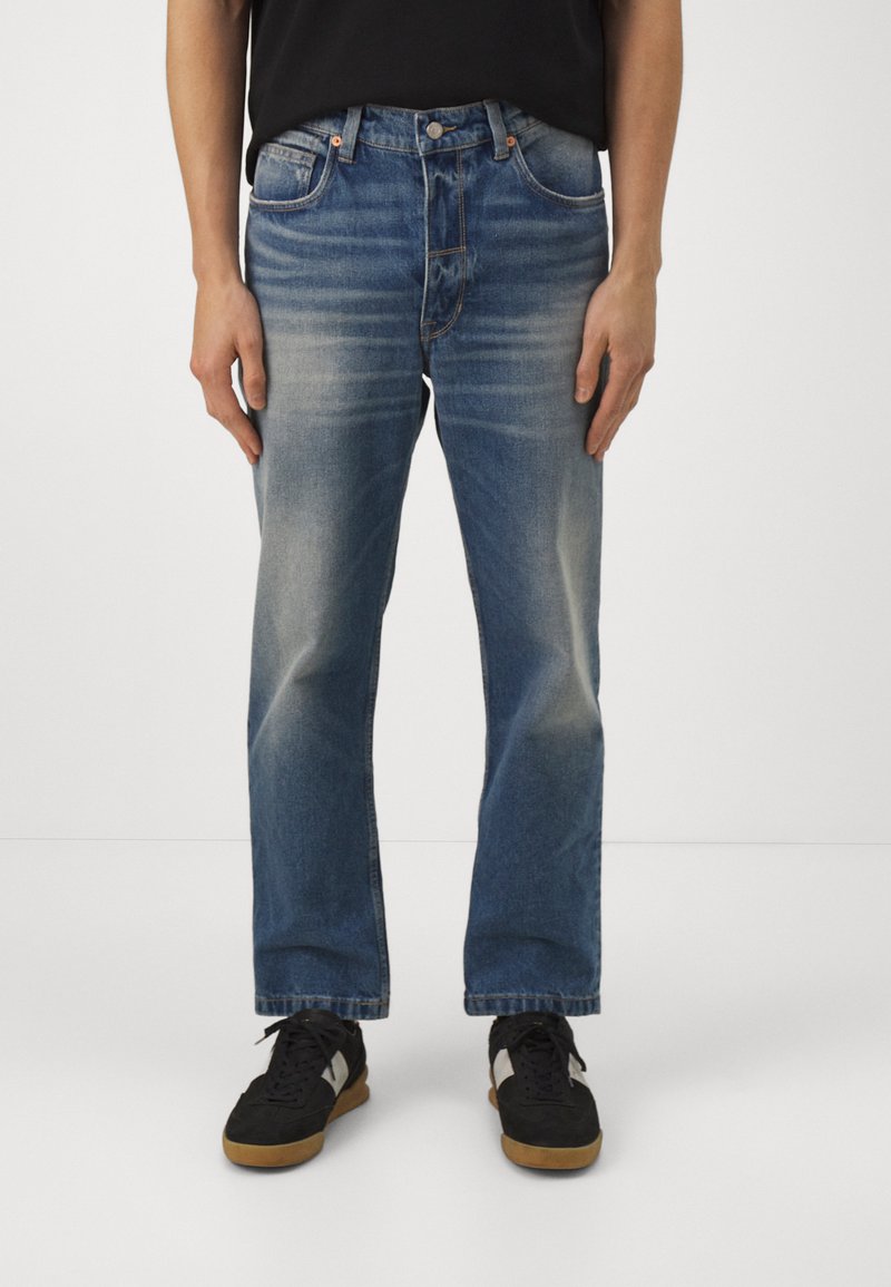 Vaqueros de mezclilla azul con un acabado desteñido, diseño de pierna recta, estilo de cinco bolsillos y herrajes de metal. Llevados con zapatillas negras.