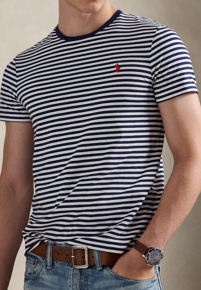 Άνδρας με μπλε-ναυτικό και λευκή ριγέ μπλούζα με κόκκινο λογότυπο, καφέ δερμάτινη ζώνη, μπλε τζιν και ρολόι χειρός με καφέ λουράκι, με το χέρι στην τσέπη.