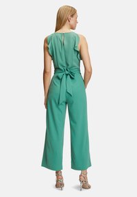 Grön jumpsuit med volangkrage, knytliv, vida ben och en nyckelhålsdetalj på baksidan. Har slät tyg och inga synliga mönster.