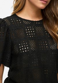 Blouse noire texturée avec un motif en damier, dotée de manches courtes à volants et de détails en tissage ouvert, mettant en valeur un design léger et aéré.