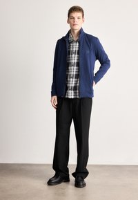 Nāvija zilas zipā uzvelkams džemperis pār melnu un baltu rūtainu kreklu, kombinēts ar melnām biksēm un melniem apaviem, uz neitrālas fona.