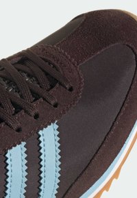 Brązowy sneaker wykonany z połączenia zamszu i materiałów tekstylnych, z jasnoniebieskimi zygzakowymi paskami i podeszwą z gumy.