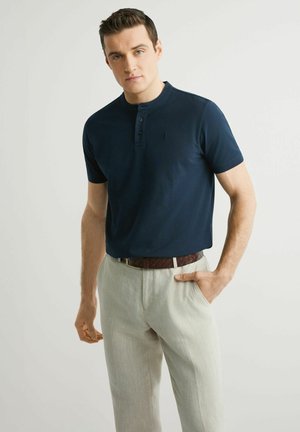 Mężczyzna w granatowej koszulce henley z krótkim rękawem, wpuszczonej w jasnobeżowe spodnie z brązowym plecionym paskiem, stojący z jedną ręką w kieszeni.