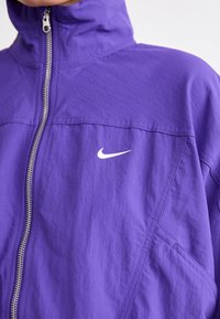 Nike purpurinė striukė su aukštu apykakle ir pilnu užtrauktuku. Yra balta logo, tekstūruota medžiaga ir matomos siuvimo detalės.