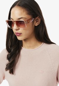 Översized solglasögon med en klar, gradient rosa ram och brunt tonade linser; bärs med en ljusrosa stickad tröja.