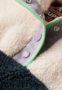 Veste en polaire avec une texture crémeuse et duveteuse, une section inférieure bleu marine, et des boutons-pression violets avec une bordure verte le long du col.