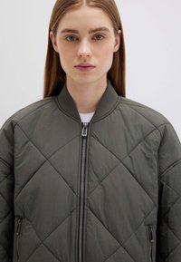 Giacca bomber trapuntata verde scuro con collo rotondo, chiusura frontale con cerniera e tasche laterali, realizzata in tessuto leggero.