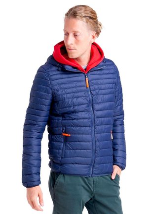 Un homme regarde sur le côté, vêtu d’une veste matelassée bleu marine à capuche rouge et d’un pantalon vert foncé, sur fond blanc.