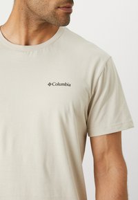 T-shirt en coton beige clair avec un col rond et des manches courtes, portant un petit logo noir Columbia sur la zone de la poitrine.