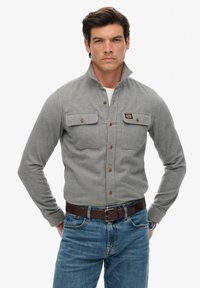Graues Hemd mit Stehkragen, zwei Brusttaschen und braunen Knöpfen; getragen mit blauen Jeans und einem braunen Gürtel.