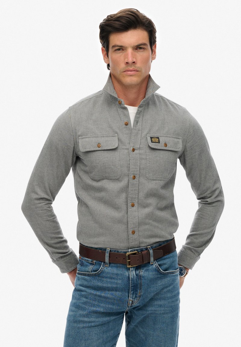 Graues Hemd mit Stehkragen, zwei Brusttaschen und braunen Knöpfen; getragen mit blauen Jeans und einem braunen Gürtel.