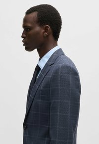 Giacca da abito a quadri blu scuro con revers a punta, chiusura con un solo bottone e taschino sul petto. Abbinata a una camicia blu chiaro e una cravatta scura.