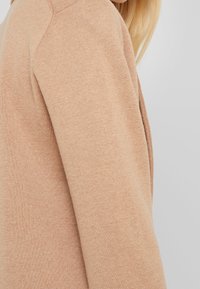 Beige gestrickter Pullover mit strukturiertem Finish, ausgestattet mit Raglanärmeln und einem subtilen, glatten Stoffmuster. Nahaufnahme des Schulterbereichs.