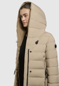 Beige pufferjakke med en stor hætte, quiltet struktur, frontlynlås, sidelommer og sorte dekorative knapper.