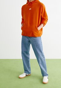 Person som bär en orange Nike-hoodie, ljusblå jeans och vita sneakers med svart Nike-swoosh, står på grön golv mot en vit vägg.