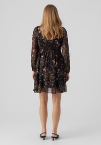 Vestido negro con flores, de mangas largas, con cintura fruncida y falda con capas. Presenta patrones florales en rosa y verde sobre un tejido transparente.