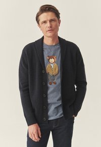 Tumši zils adīts džemperis ar šalli, piecu pogu aizdare. Gaiši pelēka T-krekls ar lāča grafiku, kas valkā džemperi un bikses.