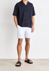 Camisa de manga corta en navy con botones, pantalones cortos a rayas claros y sandalias negras de tipo deslizante. Ajuste casual, tejido suave y diseño relajado.