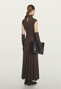 Robe marron sans manches avec un dos à ouverture en forme de goutte, associée à de longs gants en cuir noirs et un sac noir. Texture lisse et design fluide.