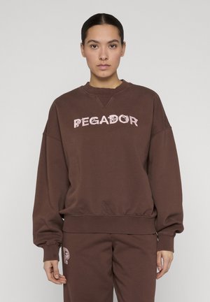 Donna che indossa una felpa oversize marrone e pantaloni abbinati con la scritta "PEGADOR" in rosa chiaro sul petto e sui pantaloni.