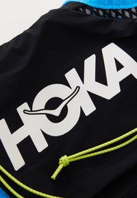 Black fabric sports gear with a large white "HOKA" logo. Features bright blue accents and a yellow drawstring with a textured finish.