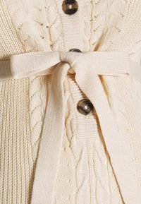 Cardigan en tricot torsadé crème avec des boutons ronds et une ceinture en tissu à la taille. Le motif de tricot texturé ajoute un intérêt visuel.