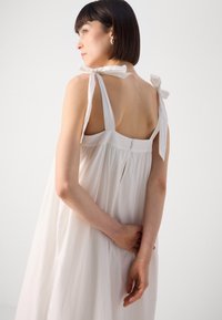 Vestido blanco, ligero y con un corte holgado; cuenta con tirantes gruesos atados en forma de lazo, una espalda con abertura en forma de llave y pequeños detalles de botones.
