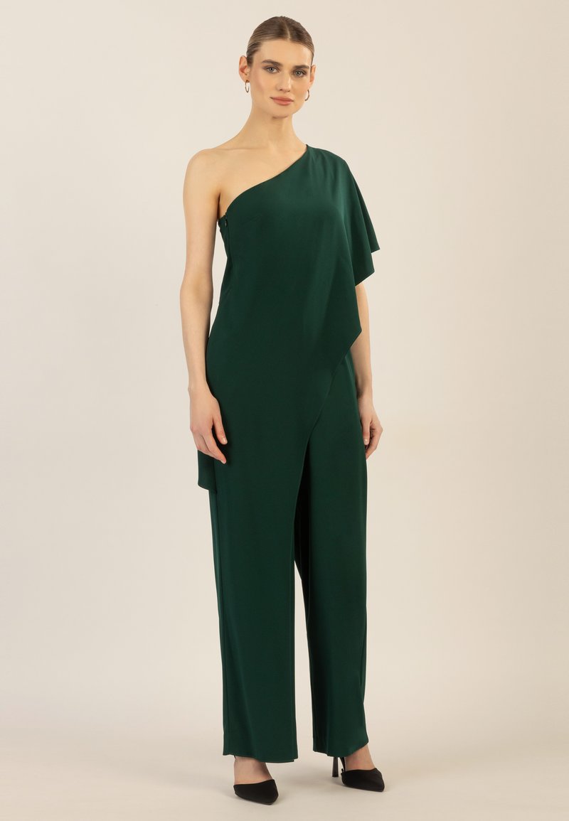 Grøn one-shoulder jumpsuit med asymmetrisk drapering, lavet af glat stof, med brede ben og sorte spidse hæle.