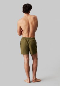 Pantaloni da bagno verdi con una cintura elastica, tasca sul retro e una superficie liscia, indossati da una persona con il busto nudo.