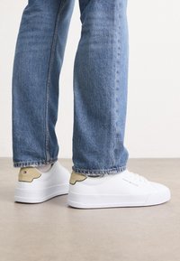Weiße Sneakers mit glatter Textur, verziert mit einem beigen Akzent an der Ferse und einem Logo. Kombiniert mit hellblauen Denim-Jeans.