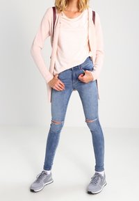 Cardigan rosa claro sobre una blusa blanca, combinado con jeans desgastados azules y zapatillas grises. El atuendo presenta estilos casuales y ajustados, con texturas en capas.