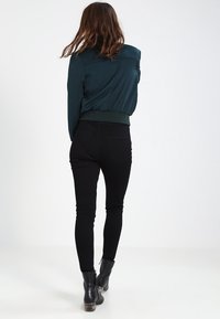 Žena s hnědými vlasy, oblečená v tmavě zelené bundě, černých skinny džínách a černých kotníkových botách, odchází na bílém pozadí.