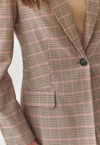 Blazer à carreaux marron avec un col cranté, fermeture à un bouton et subtils accents rose dans le motif du tissu.