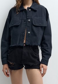 Przycięta czarna kurtka dżinsowa z dwoma kieszeniami na piersi, srebrnymi zatrzaskami i frędzlami na dole. W połączeniu z czarnymi, przetartymi szortami.