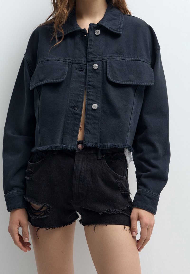 Przycięta czarna kurtka dżinsowa z dwoma kieszeniami na piersi, srebrnymi zatrzaskami i frędzlami na dole. W połączeniu z czarnymi, przetartymi szortami.