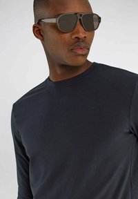 Maglietta nera a maniche lunghe con scollo tondo e un logo sottile. La persona indossa occhiali da sole scuri con un design geometrico e lenti specchiate.