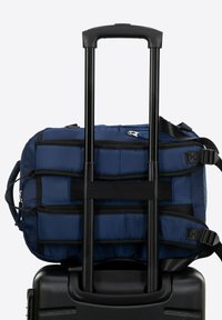 Námořnická modrá cestovní taška na sportovní vybavení připevněná na černém kufru na kolečkách. Taška má polstrovaný design, popruhy a zip. Kufr má hladký, texturovaný povrch.