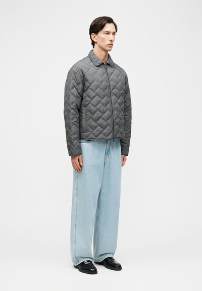 Άνδρας που φοράει γκρι μπουφάν με φερμουάρ και καπιτονέ σχέδιο, ανοιχτό μπλε τζιν παντελόνι με φαρδιά πόδια και μαύρα παπούτσια, στέκεται μπροστά σε ένα απλό λευκό φόντο.