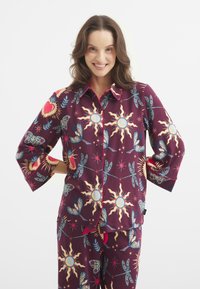 Paarse pyjama met een patroon van zonnen, harten en vogels. Korte mouwen met contrasterende rode bies en een ontspannen pasvorm.