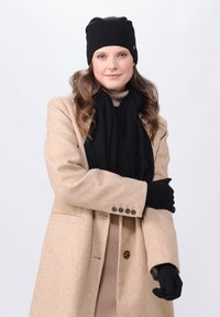 Berretto di maglia nero, sciarpa nera, guanti neri e cappotto beige con dettaglio a tre bottoni; il modello si trova di fronte a uno sfondo bianco uniforme.