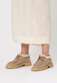 Stivaletti beige in suede con una morbida fodera color crema, dotati di una suola spessa e lacci marroni, posizionati su uno sfondo chiaro.