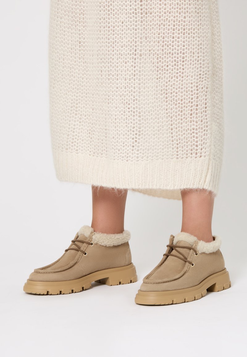 Stivaletti beige in suede con una morbida fodera color crema, dotati di una suola spessa e lacci marroni, posizionati su uno sfondo chiaro.