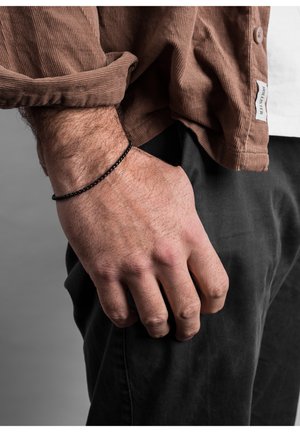 Braccialetto di perline nere al polso, vista ravvicinata. Mano appoggiata su pantaloni scuri, con la manica di una camicia di velluto a coste marrone arrotolata.