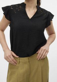 Czarna bluzka o teksturowanym wykończeniu, z dekoltem w kształcie litery V, z koronkowymi detalami w kwiaty na ramionach oraz krótkimi falbaniastymi rękawami, połączona z beżowymi spodniami.