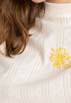 Cheveux bruns reposant sur un pull en maille crème avec des motifs délicats et une fleur jaune brodée près de l'épaule.